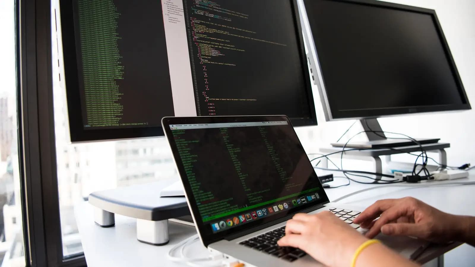 Developer typing code on a laptop in front of several monitors full of code